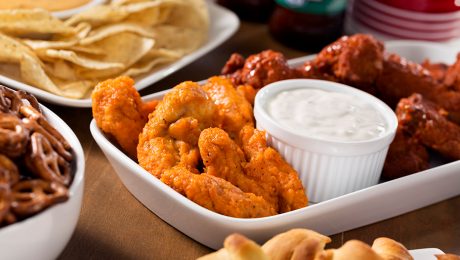 Buffalo wings in two flavours on a plate with a bowl of creamy sauce and other party snacks on a brown table