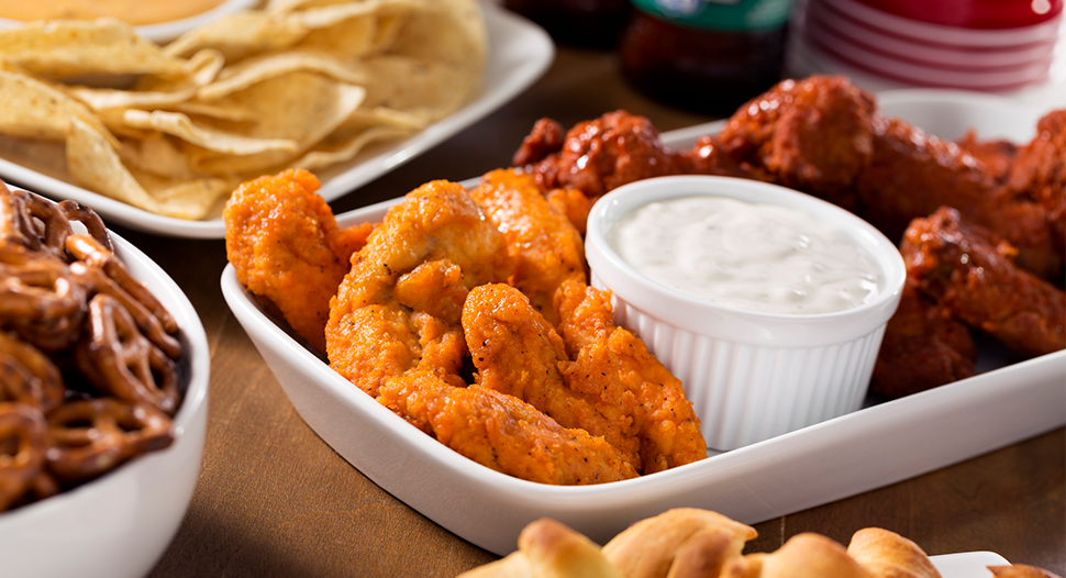 Buffalo wings in two flavours on a plate with a bowl of creamy sauce and other party snacks on a brown table