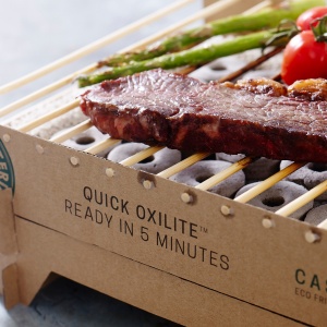 A thin slab of steak, two sticks of asparagus and tomatoes on the vine being cooked on a portable BBQ grill made of cardboard and bamboo